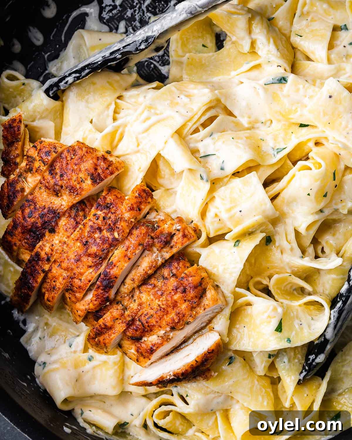 Chicken alfredo pasta in black pan with tongs.