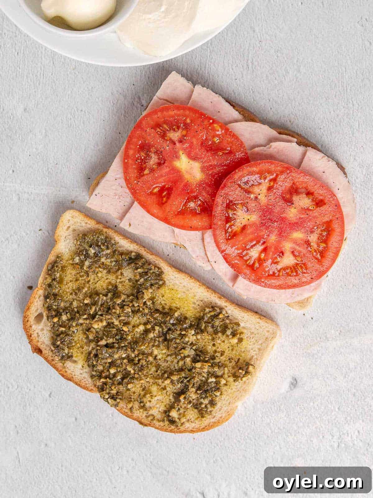 Grilled Turkey Pesto and Mozzarella Sandwich Recipe 6 Two pieces of sourdough bread with pesto layered across the top. One one slice of bread, thick slices of turkey are layered on top with tomato slices on top of the turkey. The tomato slices have been salted and peppered.