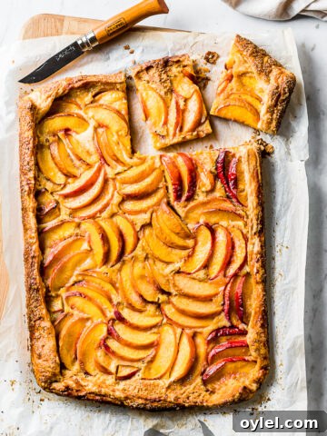 A golden brown rectangular peach galette rests atop white parchment paper on a wooden cutting board. Two squares are cut out of the top right corner with a wooden handled paring knife.