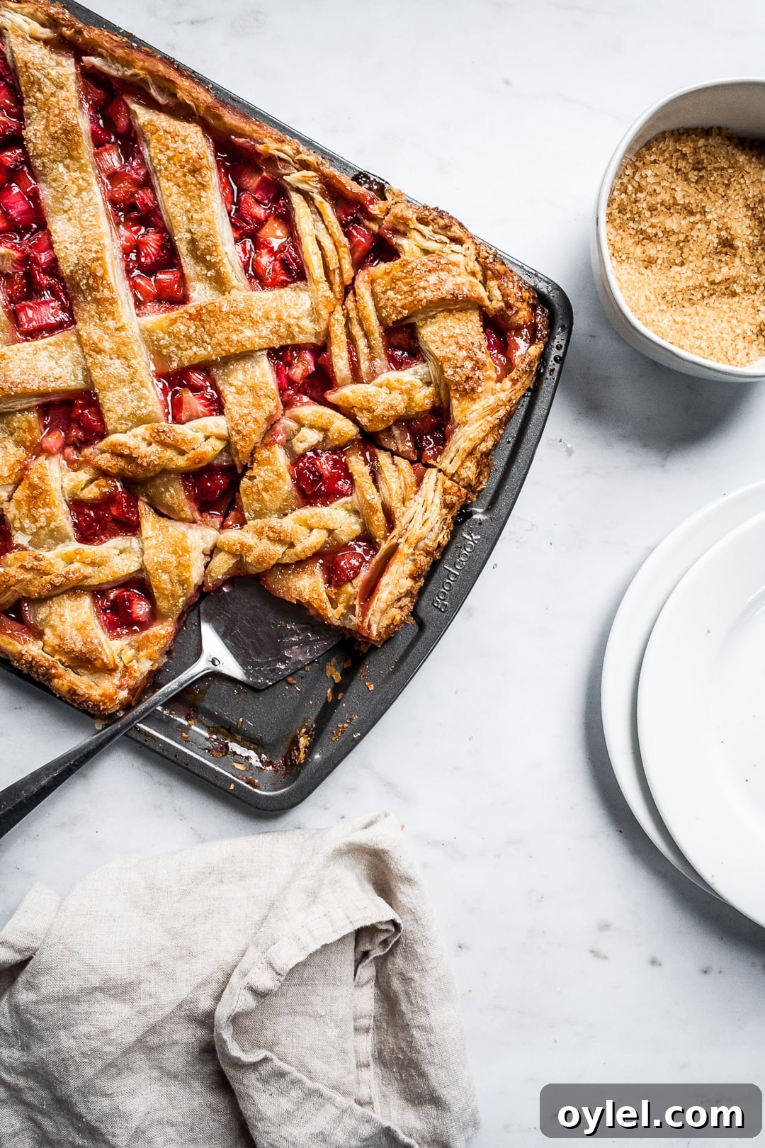 Baked strawberry rhubarb slab pie in a sheet pan with several cut slices and a serving utensil on a marble background