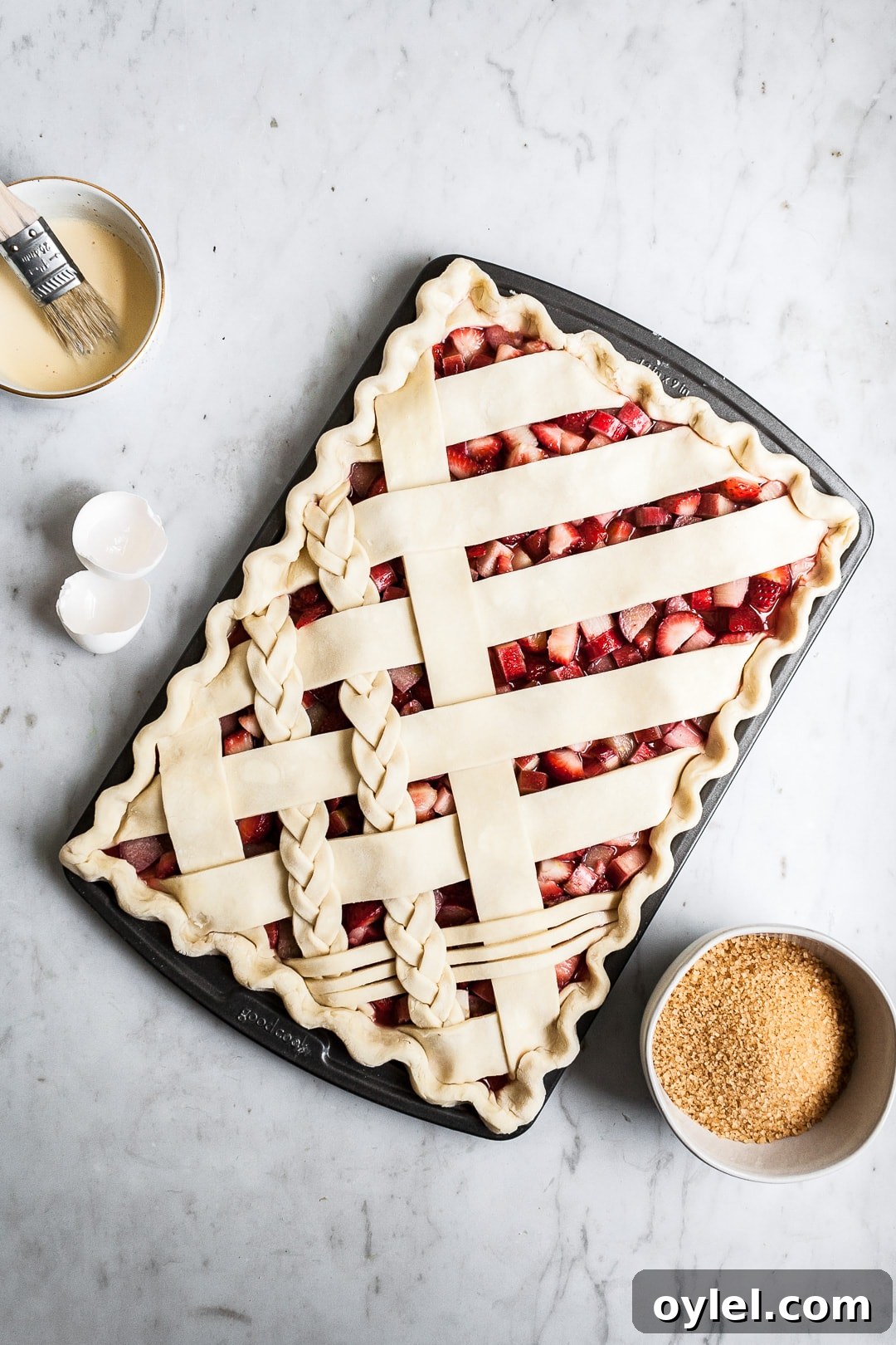 Crimped unbaked lattice slab pie crust in sheet pan on a marble background with bowls of sugar and egg wash nearby
