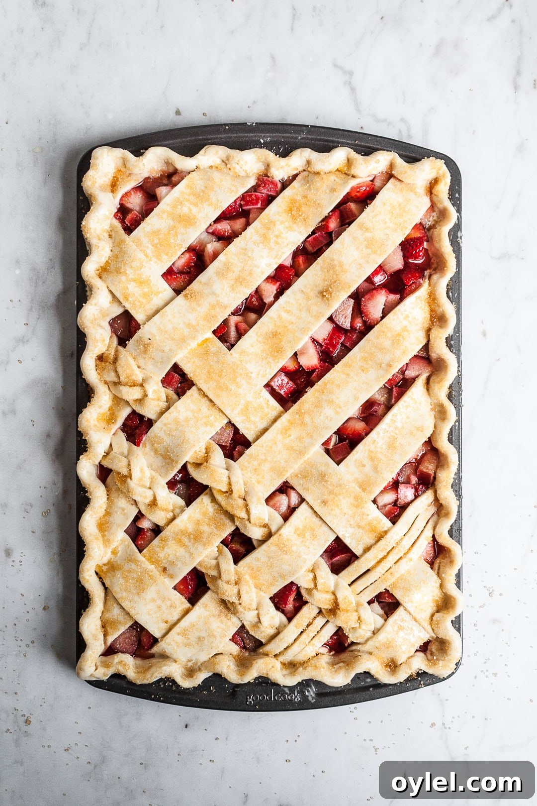 Top view of unbaked crimped lattice slab pie with strawberry rhubarb filling, sprinkled with coarse sugar, on a marble background