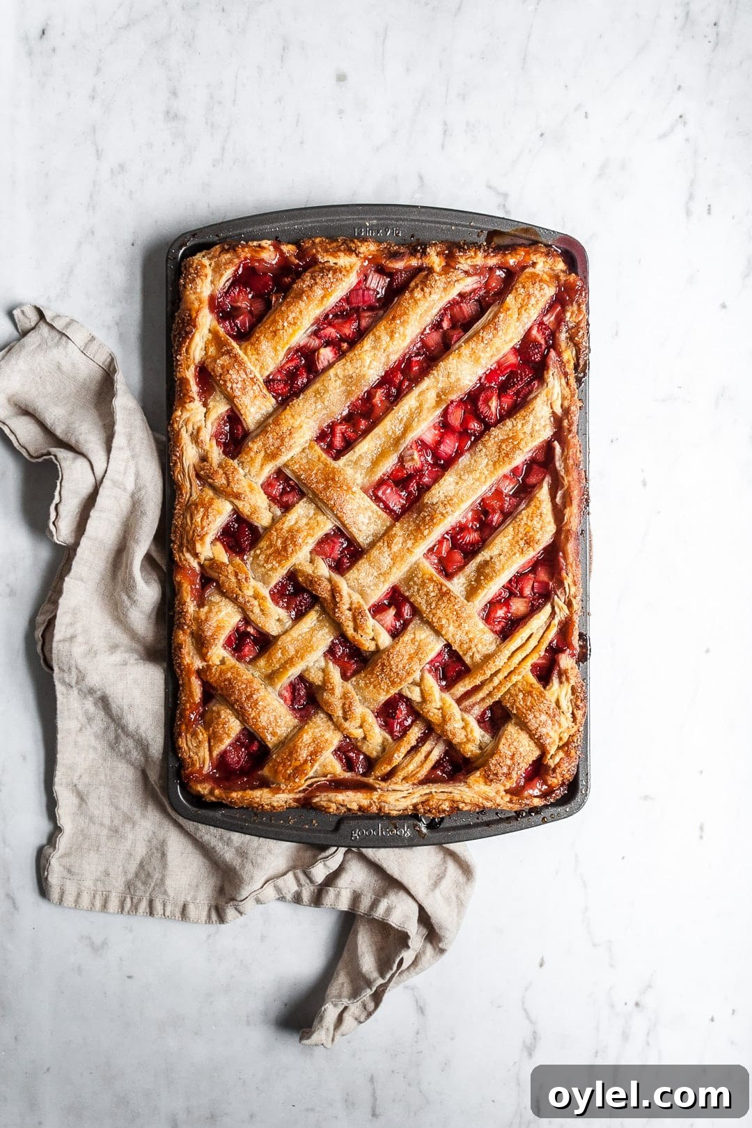 Top view of baked strawberry rhubarb slab pie with natural linen towel underneath pan, resting on a marble background