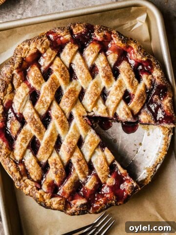 A golden brown lattice topped baked cherry rhubarb pie on a parchment lined baking sheet. A slice has been removed from the pie.