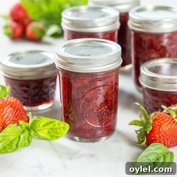 A jar of strawberry preserve sits on a table near basil and strawberries.