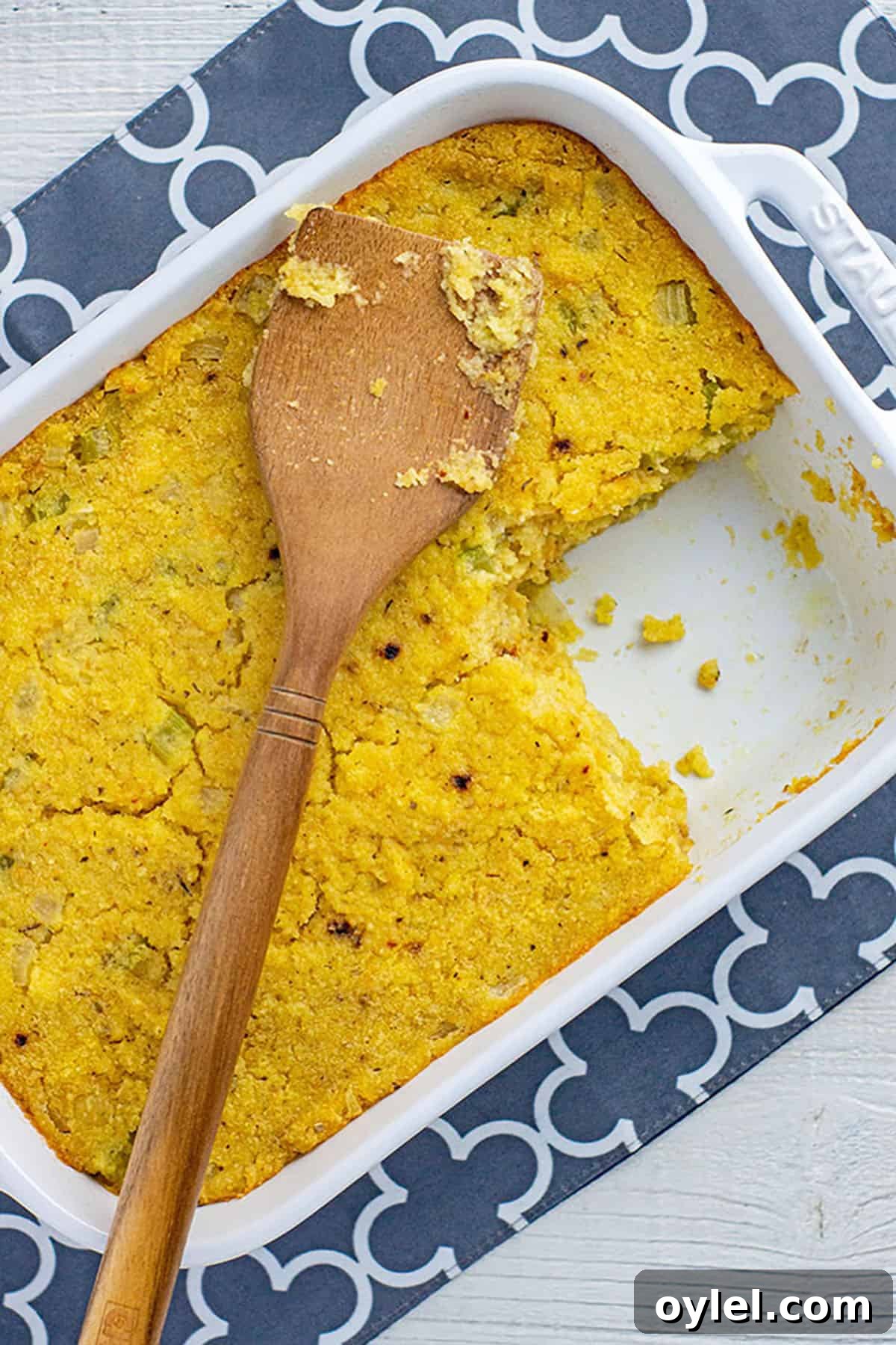 Classic Southern Cornbread Dressing Recipe 2 baked cornbread dressing in white rectangular casserole dish with wooden serving spatula on top. One serving of dressing from the corner is missing from the casserole dish.
