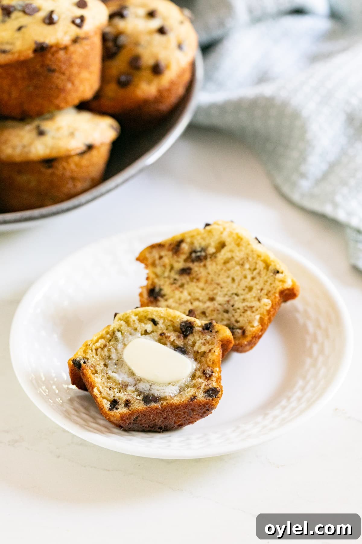 A muffin cut in half with butter melting on the front half on a white plate with other muffins behind it.