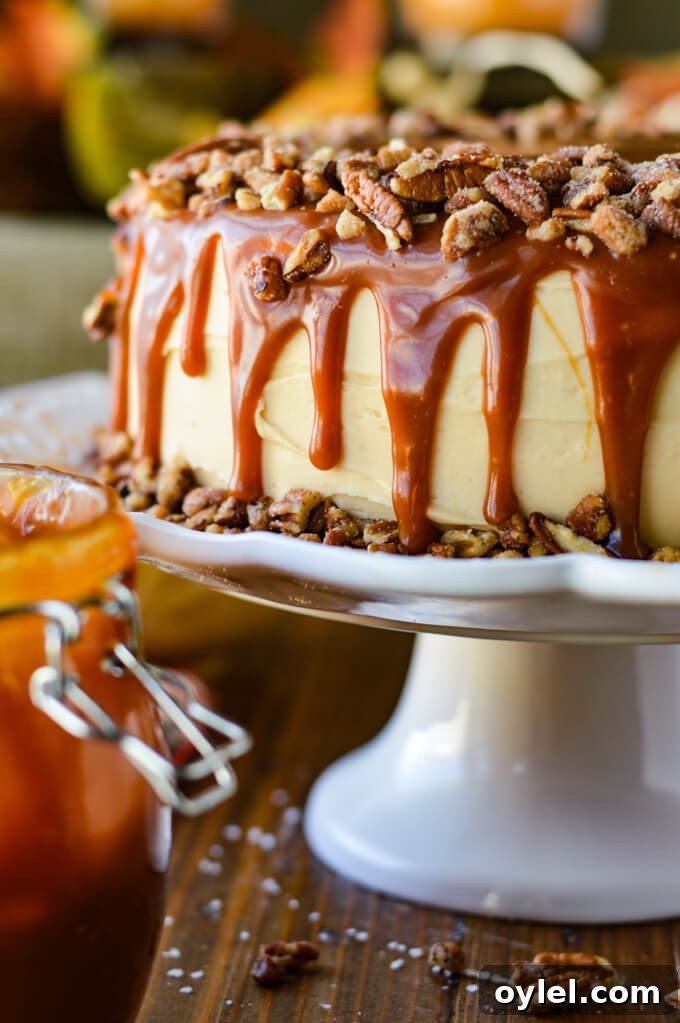 A frosted layered pumpkin cake with caramel dripping down its sides and coated on top with a layer of pecans. The cake sits on a white cake platter with a jar of caramel sauce beside it.