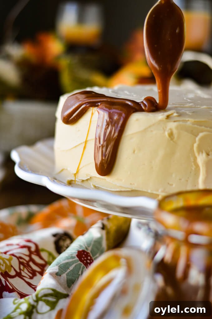A frosted pumpkin layered cake sits on a white cake stand and caramel is being drizzled on its sides.