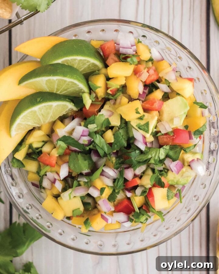 A overhead image of a bowl of fresh pico de gallo with mango with limes and mangoes as garnish.