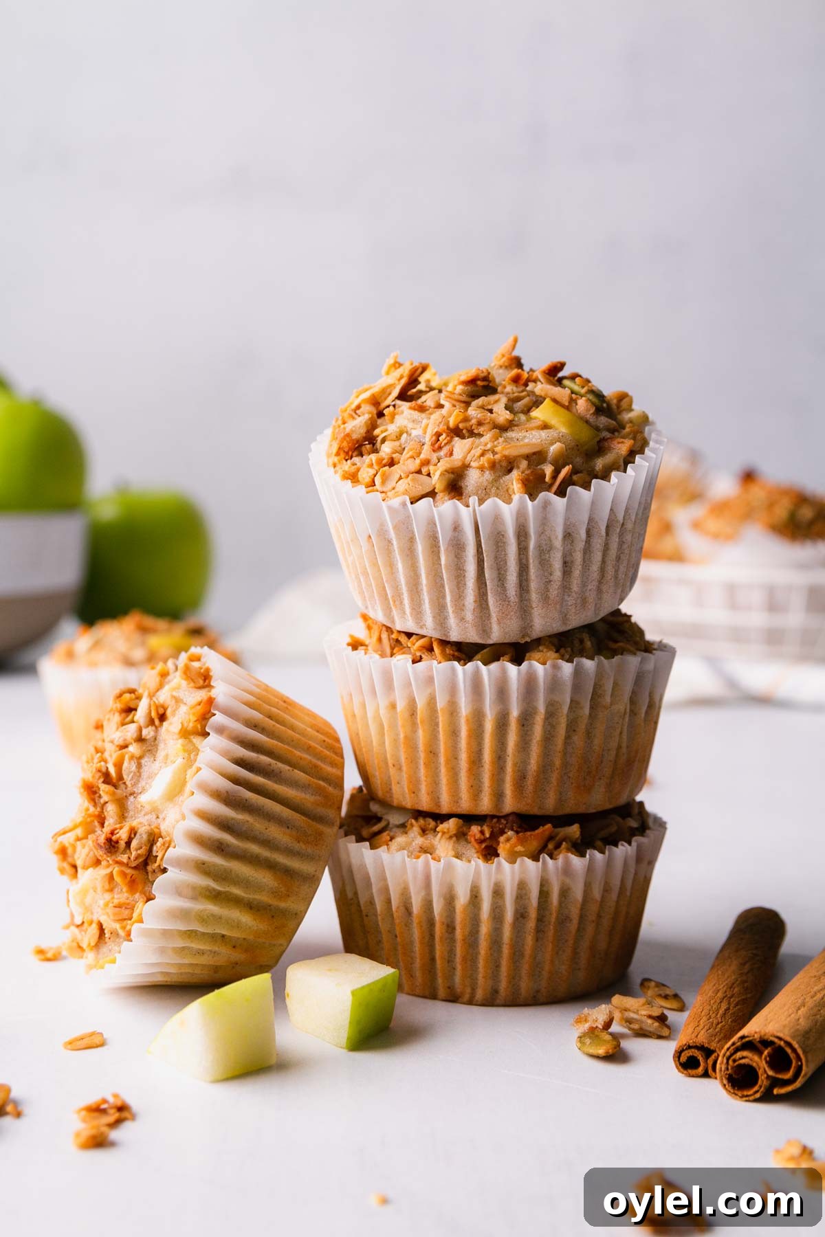 A stack of three gluten-free apple granola muffins with one more leaning on the side of the stack. Surrounded by diced apples and cinnamon sticks.