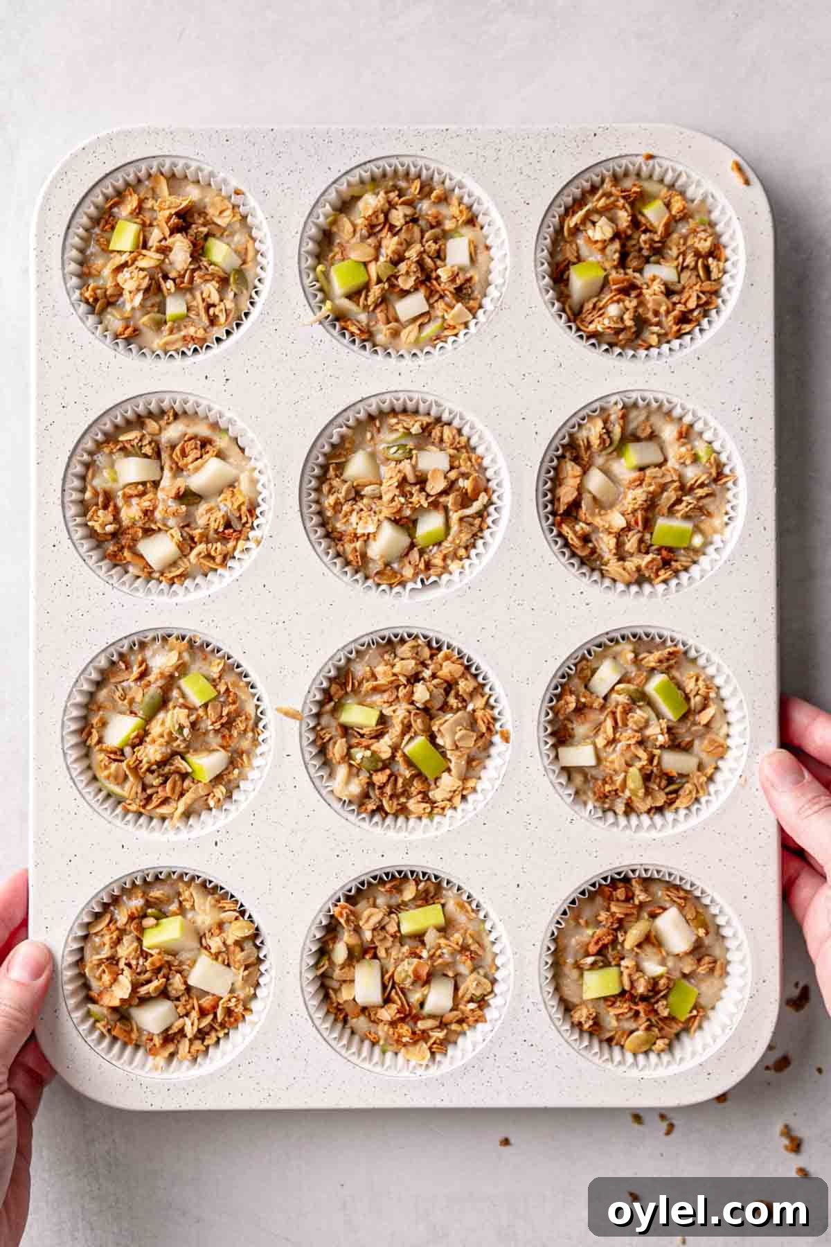 Muffin batter in white muffin tin, granola and diced apples pressed on top of batter.