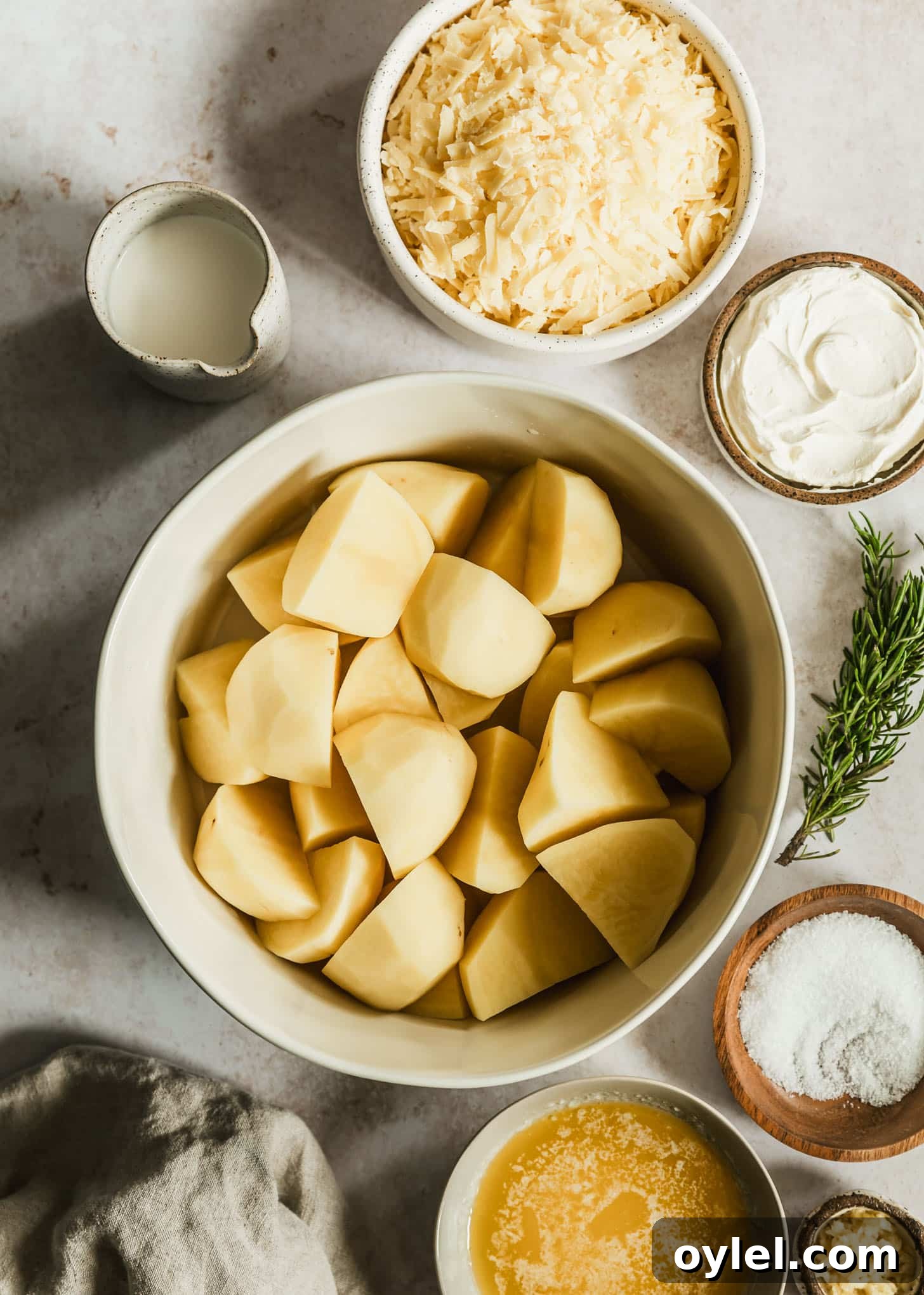 Creamy Cheesy Garlic Mashed Potatoes Recipe 2 White and brown bowls of white cheddar, potatoes, creme fraiche, cream, salt, melted butter, and garlic on a beige table next to rosemary and a tan linen.