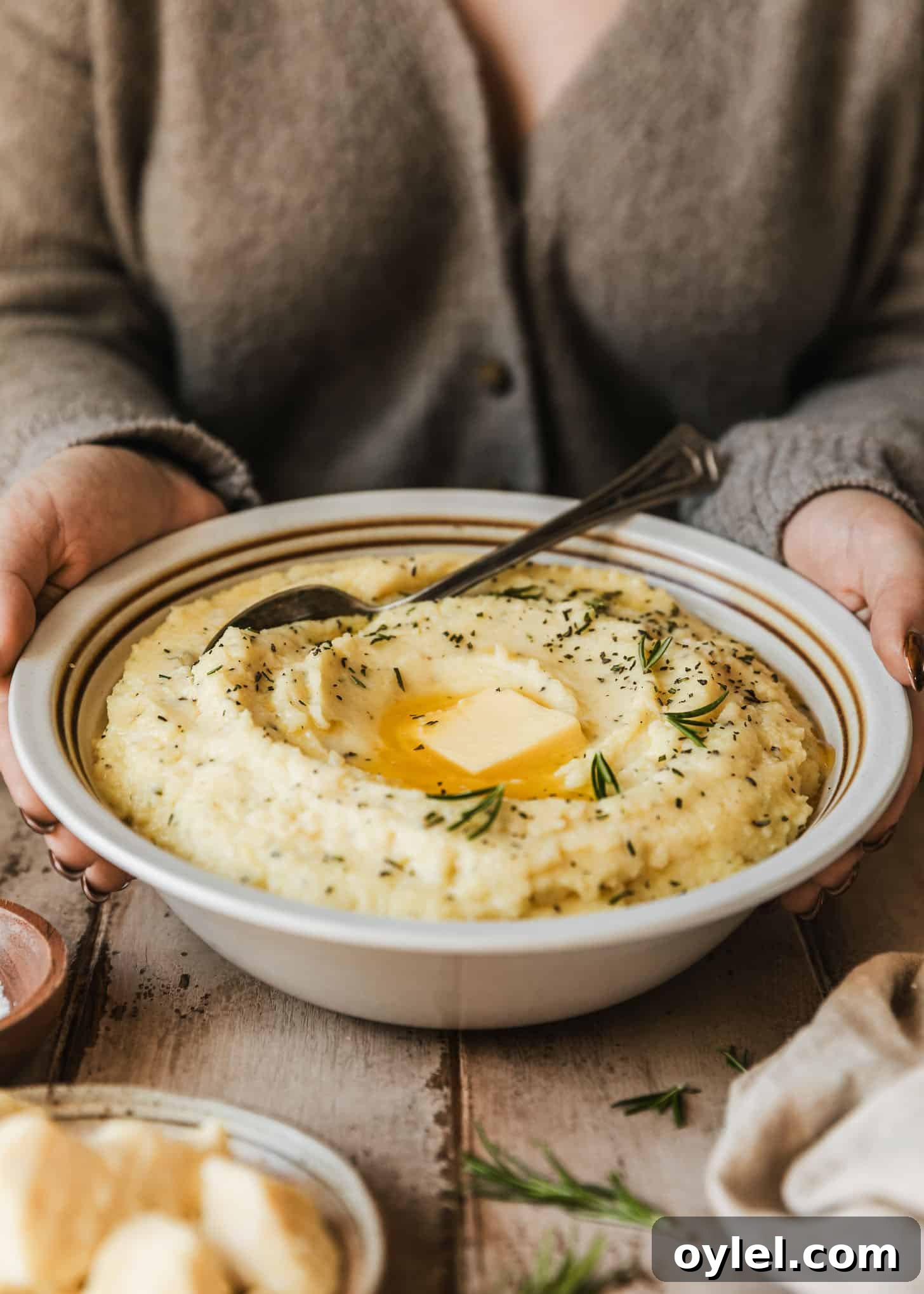 Creamy Cheesy Garlic Mashed Potatoes Recipe 5 A woman in a tan sweater holding a white bowl of cheesy garlic mashed potatoes on a wood table next to a beige linen, rosemary sprigs, and brown bowls of salt and white cheddar.