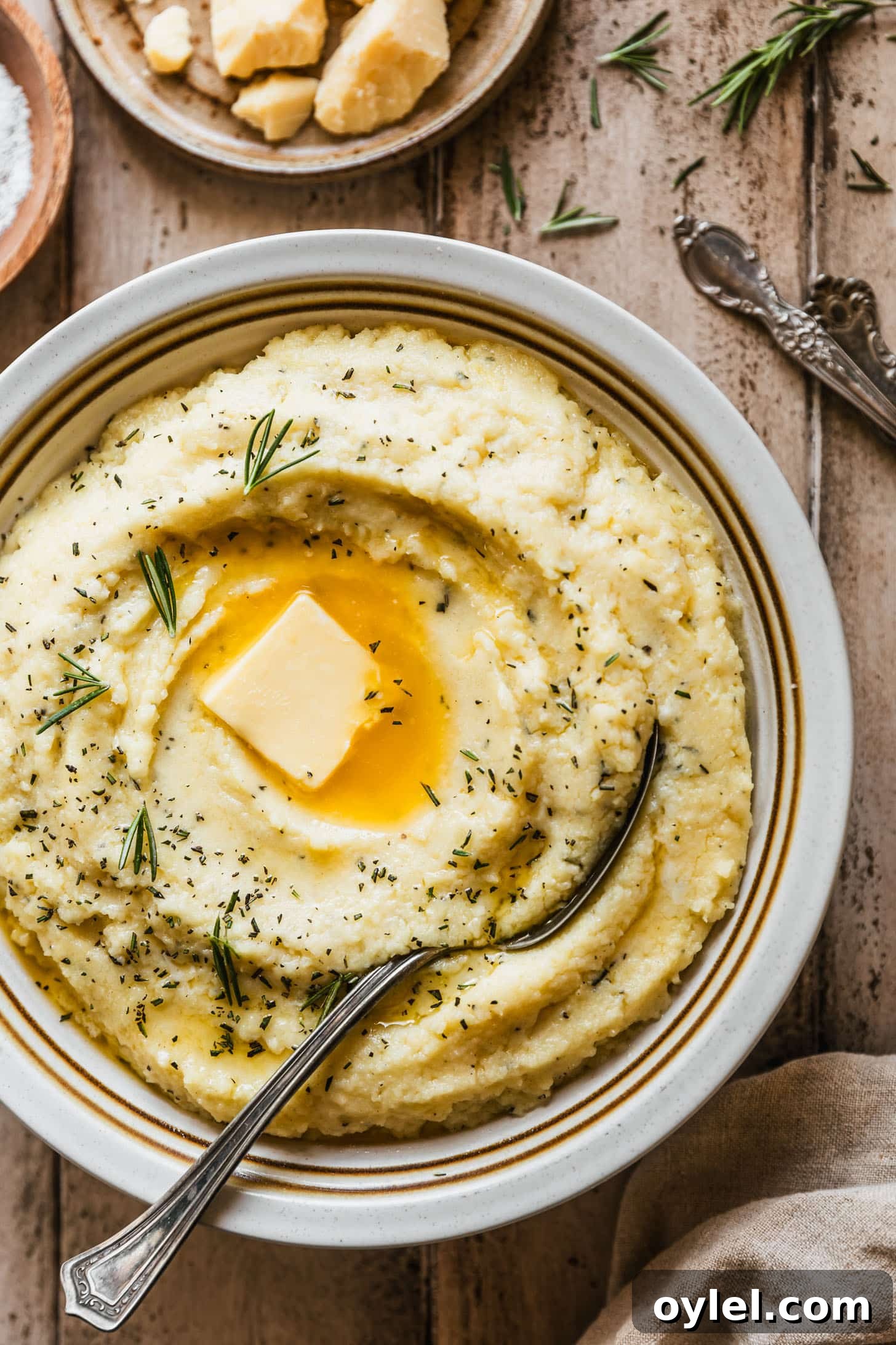 Creamy Cheesy Garlic Mashed Potatoes Recipe 6 A white bowl of garlic cheesy mashed potatoes with a spoon on a wood background next to a beige linen, rosemary sprigs, a brown plate of cheese, and vintage forks.