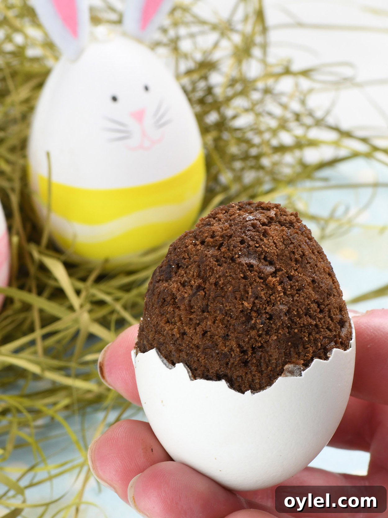 A hand holding a creative Easter dessert—a brownie baked inside a cracked eggshell. The brownie slightly overflows from the top, mimicking the look of a hatching treat. In the background, decorated Easter eggs with bunny faces and pastel colors rest on a bed of hay