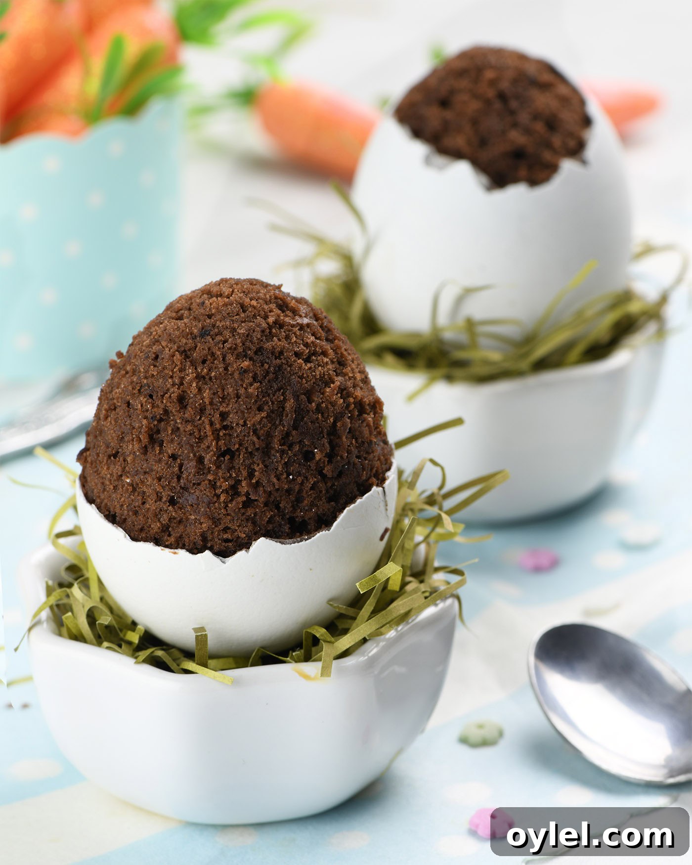 Two chocolate brownies baked inside cracked eggshells, placed on a bed of decorative grass in small white bowls, creating a festive Easter dessert presentation. A pastel background with tiny decorations and faux carrots enhances the holiday theme.