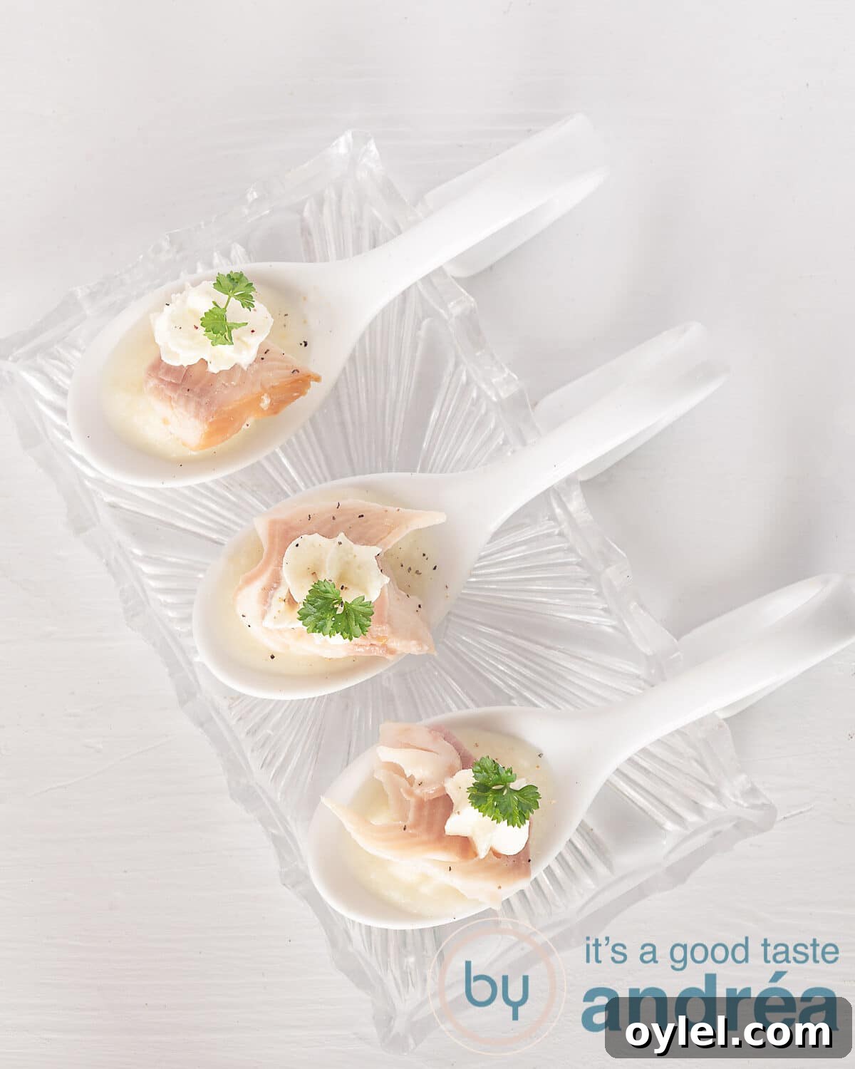 An elevated photo of three amuse-bouche spoons with asparagus puree, smoked trout and horseradish on a glass dish with a white background.