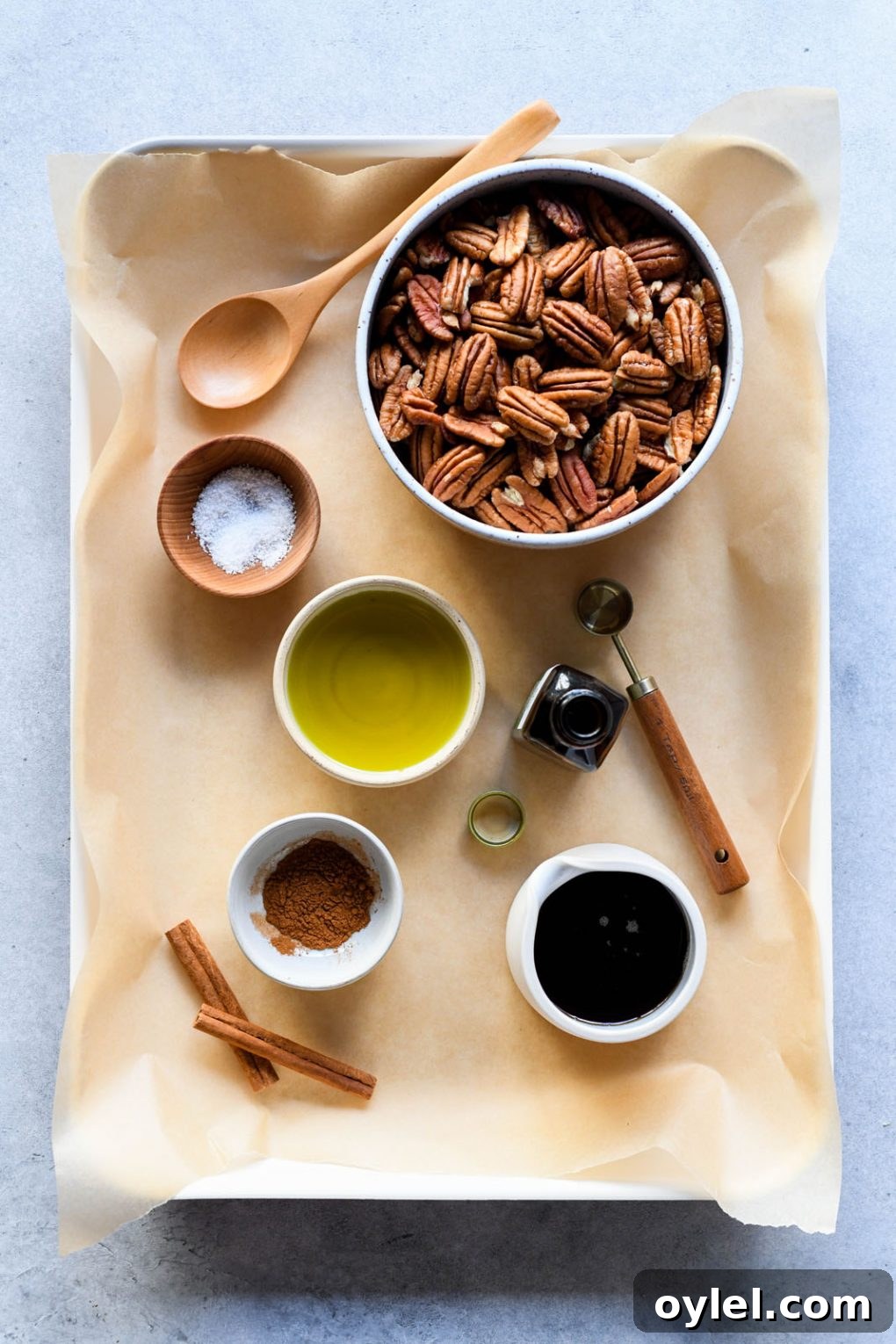 Maple Candied Pecans Recipe 3 Overhead view of a baking sheet lined with parchment paper, filled with the ingredients for candied pecans in small dishes - raw pecans, sea salt, avocado oil, cinnamon, maple syrup, and vanilla extract.