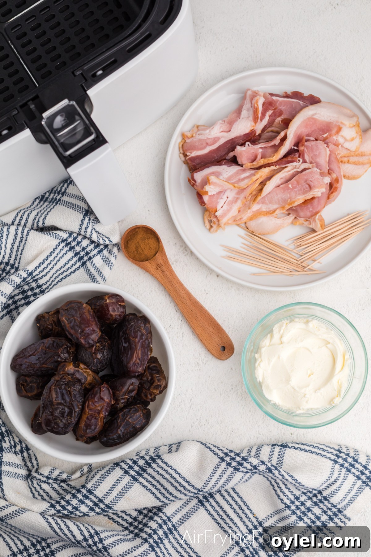 Air Fryer Bacon-Wrapped Dates Recipe 3 Table with air fryer basket, bowl of dates, cream cheese, plate with bacon and toothpicks, and a teaspoon of cinnamon.