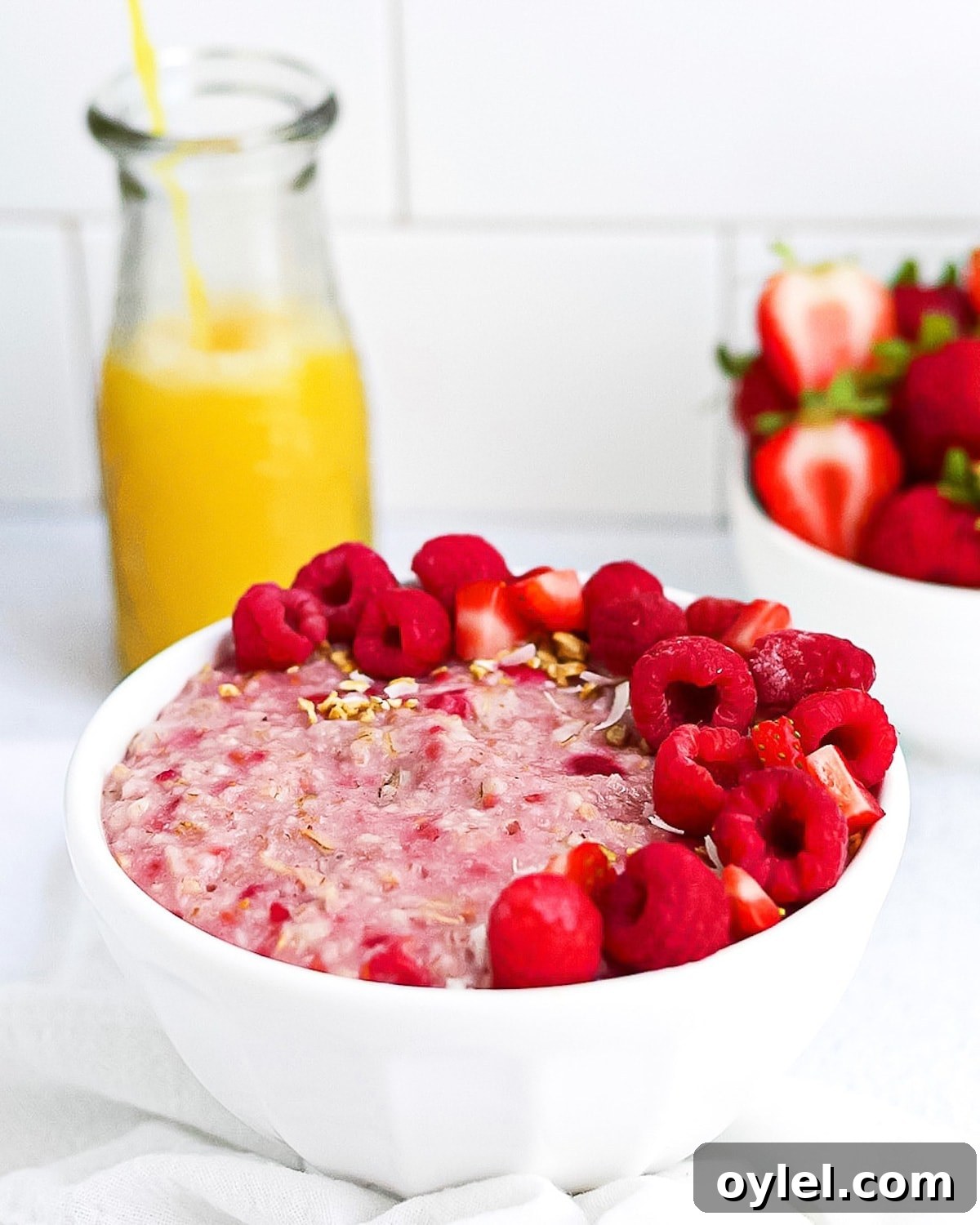 A completed picture of raspberry oatmeal garnish with fruit and granola, orange juice being poured in the background and a bowl of strawberries in the background.