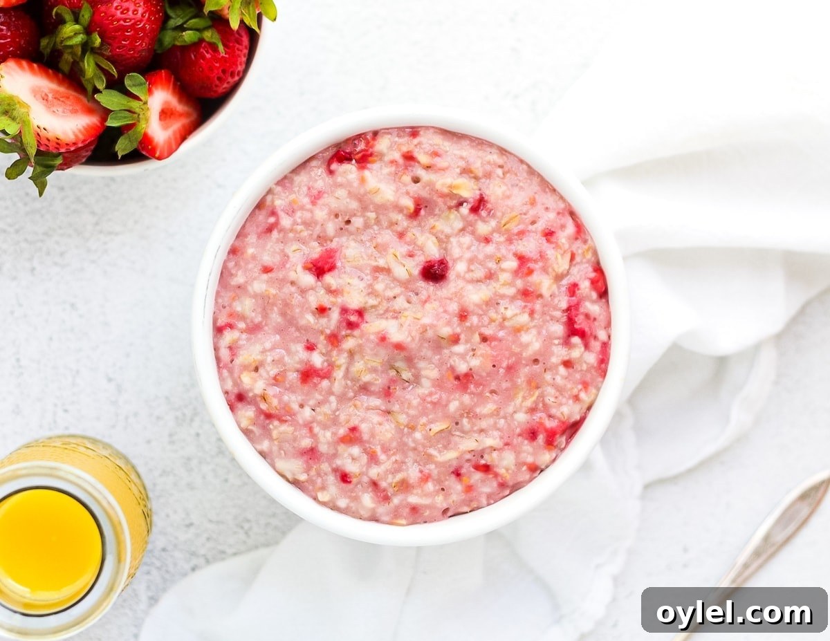 A horizontal picture containing pink raspberry oatmeal. A glass of orange juice, bowl of strawberries, a white cloth napkin, and a spoon are in the background