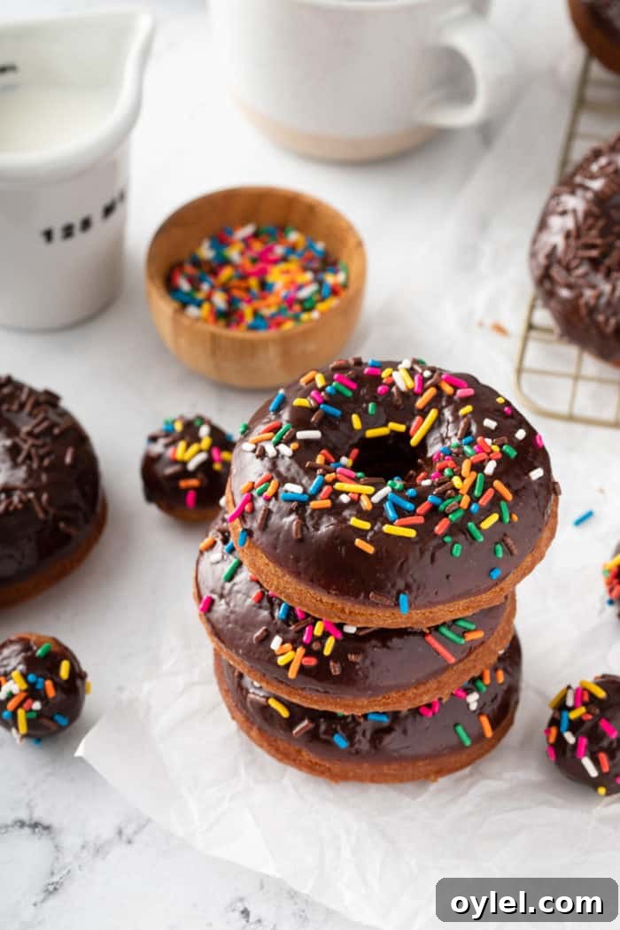 Three sprinkle-topped chocolate glazed donuts stacked on a piece of parchment paper, with more donuts and sprinkles in the background.
