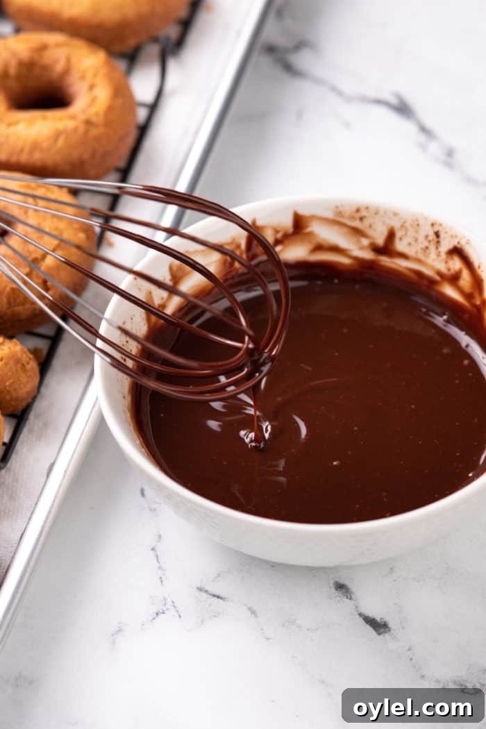 Whisk stirring chocolate glaze in a white bowl next to a pan of donuts.