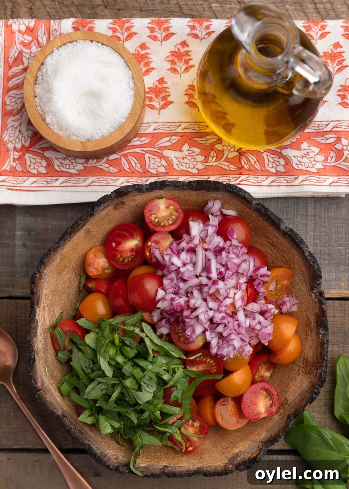 overhead photo of halved cherry tomatoes, basil, and chopped red onion in a wooden bowl, beneath a bowl of sea salt and a cruet of olive oil 