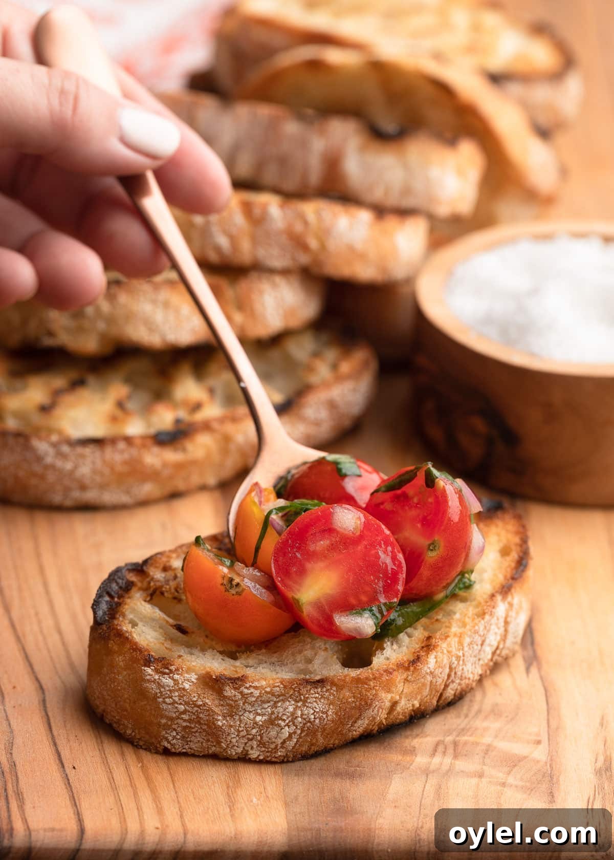 placing cherry tomato bruschetta topping onto a slice of grilled ciabatta bread with a bronze spoon on a wooden board
