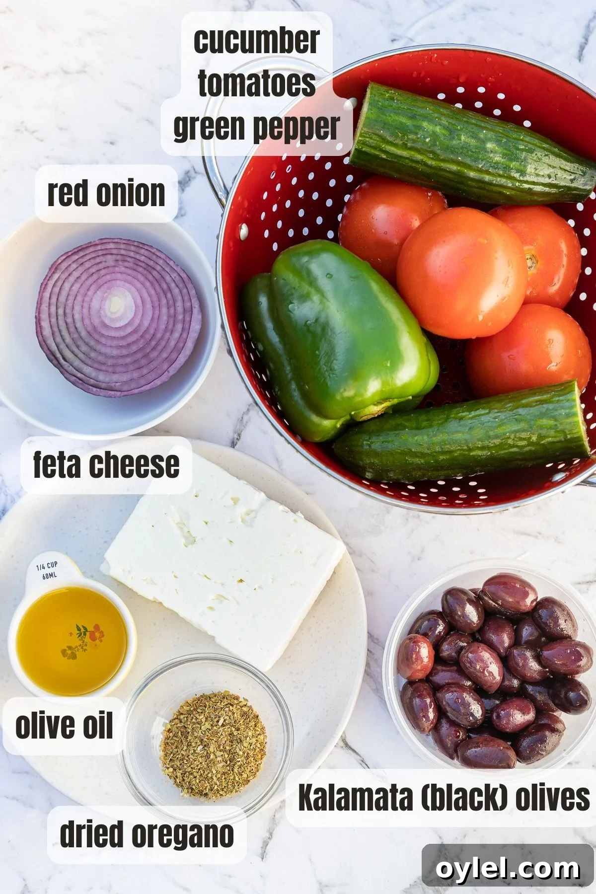 All the ingredients for a Greek salad labelled and on a marble background including tomatoes, cucumber, green peppers, dried oregano, olive oil, red onion, peppers and black olives.