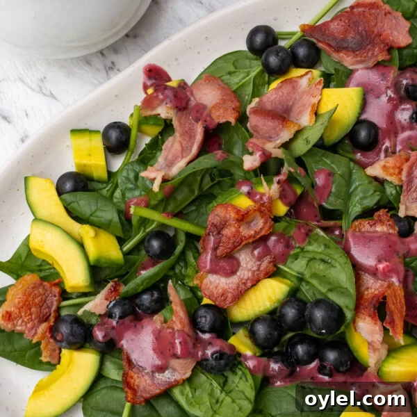An oval platter of spinach avocado salad with a white jug of blueberry dressing from above on a marble background