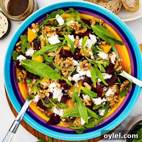 A colorful dish of beetroot and feta salad with servers in it on a wooden board and marble background with fresh mint scattered around and a jar of dressing and plate of bread at the top.