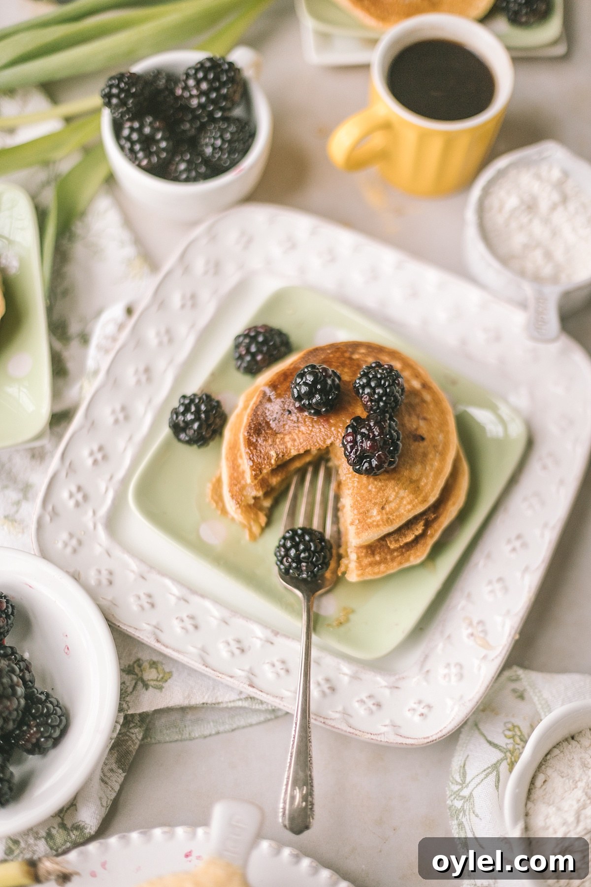 overhead shot of cornmeal pancake stack on soft green plate styled with blackberries, syrup, coffee, cornmeal