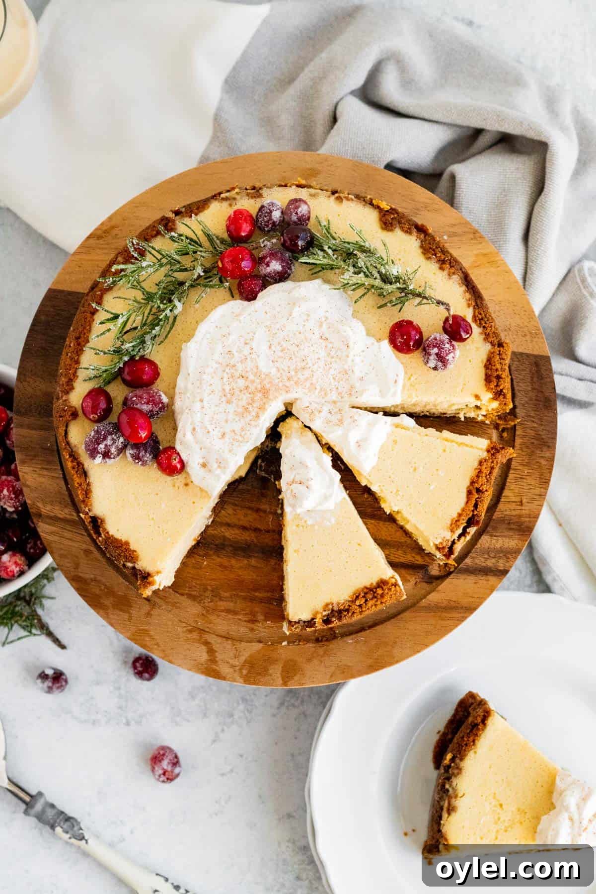 overhead view of an eggnog cheesecake decorated with cranberries and rosemary and 2 slices sliced on a wood cake stand