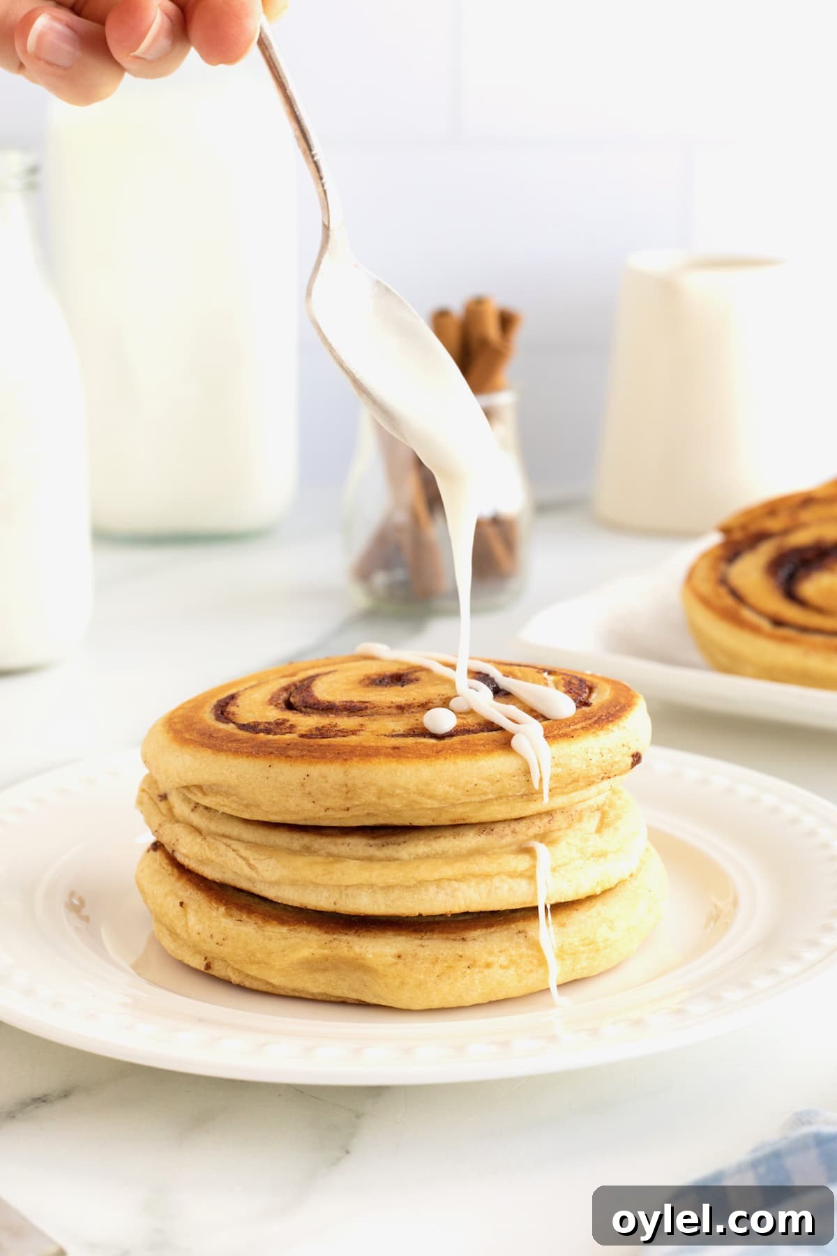 Cinnamon Roll Pancakes Recipe with Cream Cheese Glaze 2 Icing being drizzled onto a stack of three cinnamon roll pancakes on a white plate.