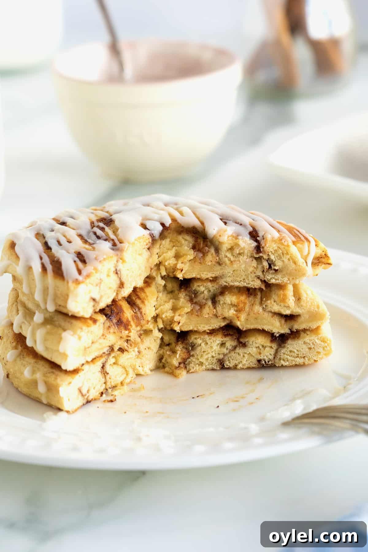 Cinnamon Roll Pancakes Recipe with Cream Cheese Glaze 4 A stack of three pancakes made out of canned cinnamon rolls on a white plate drizzled with icing.