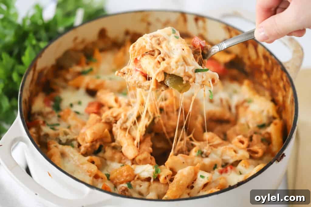 A Dutch oven full of this sausage and pepper pasta bake with a hand holding a spoon that is taking out a large scoop.