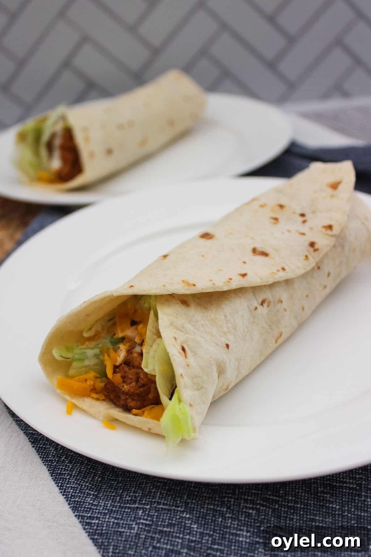 A close-up of two soft tortilla wraps filled with breaded chicken, shredded lettuce, and cheddar cheese, served on white plates with a patterned background.