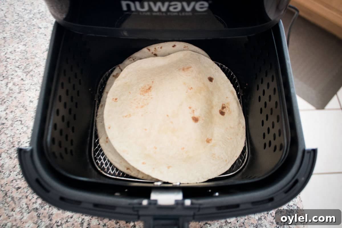 Two flour tortillas are placed inside the basket of a black air fryer on a speckled countertop, ready to be warmed.