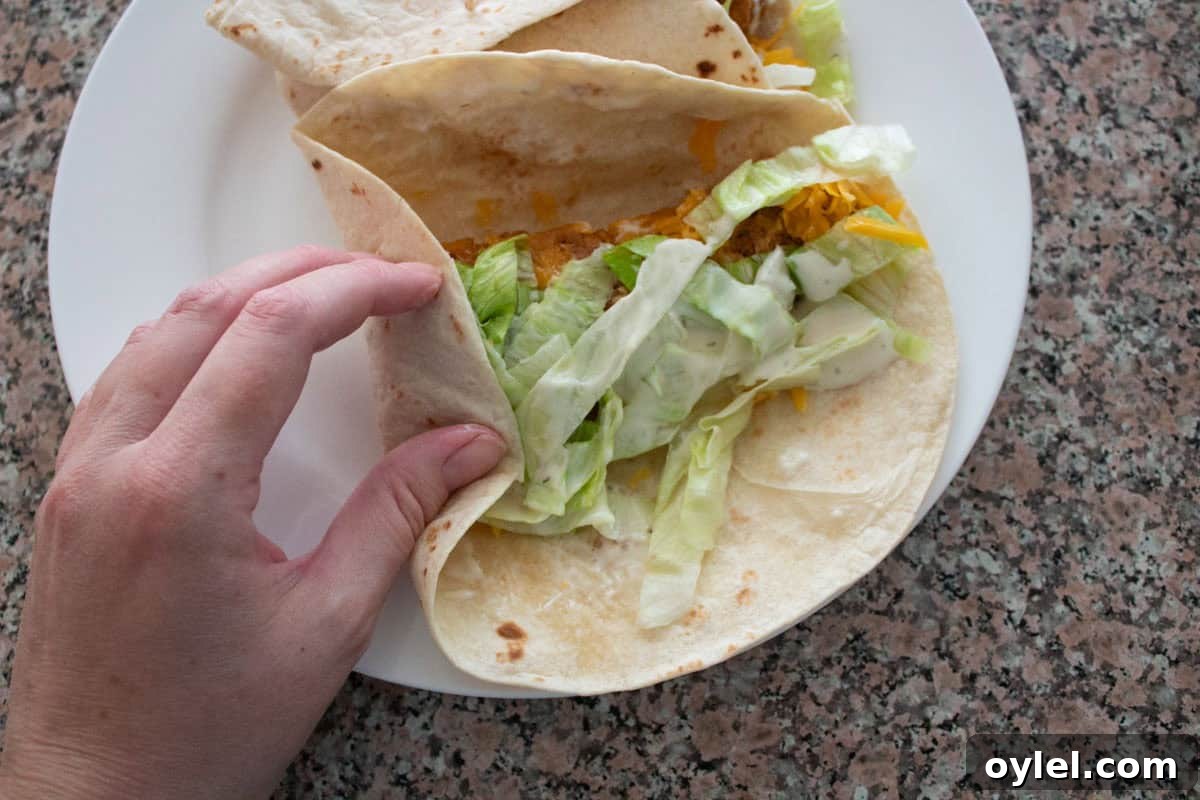 A hand holding a tortilla with lettuce, cheese, chicken, and sauce on a flour tortilla. Another wrap sits on a white plate, all on a granite countertop.