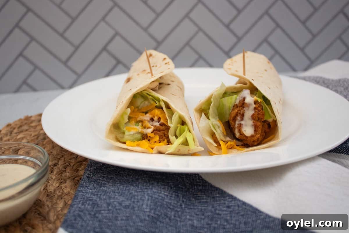 Two chicken wraps filled with lettuce, shredded cheese, and drizzled with creamy sauce, served on a white plate with toothpicks, placed on a blue and white cloth. A bowl of sauce is visible on the side.