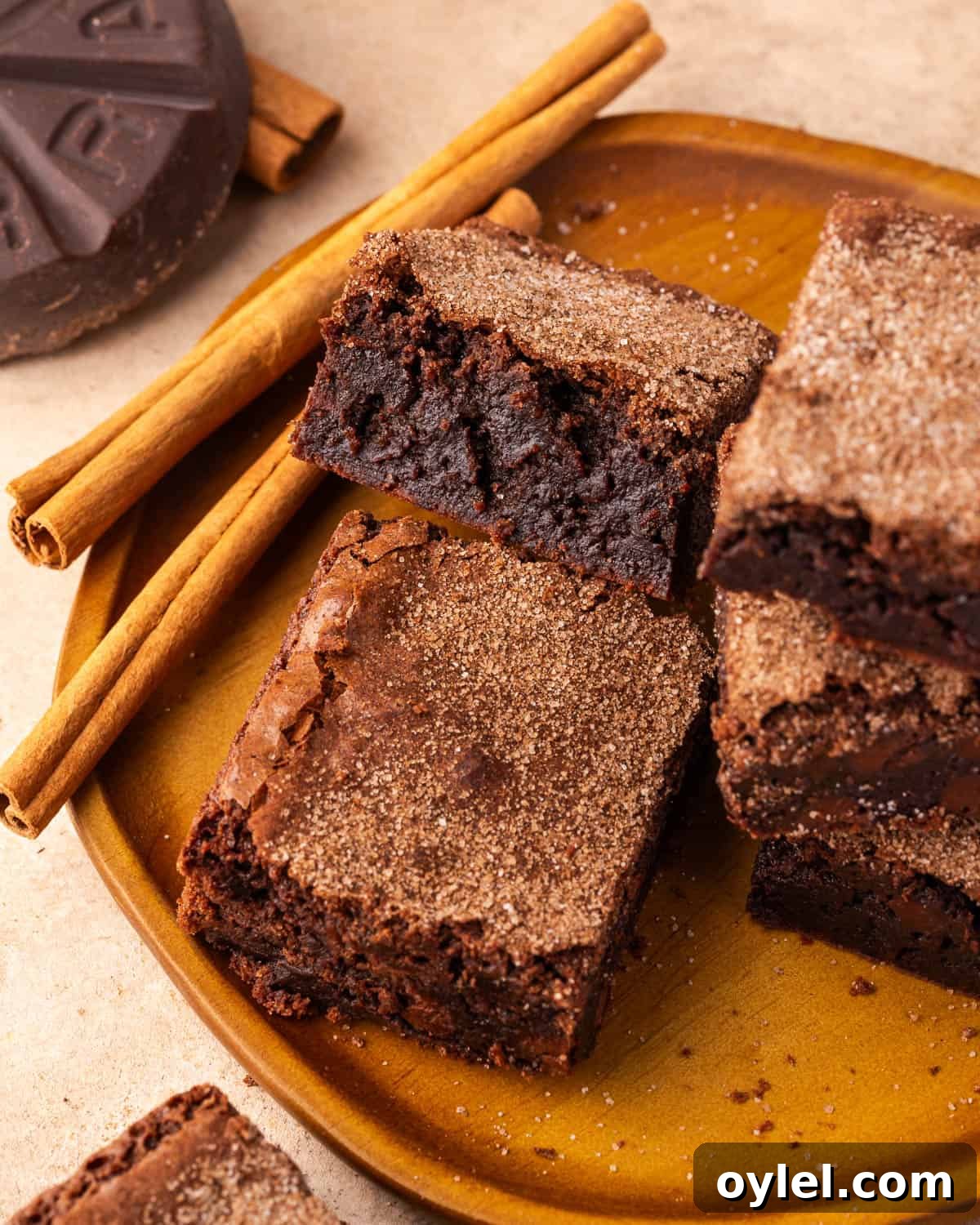 Several brownies stacked on a wood plate next to cinnamon sticks and a cocoa disc.