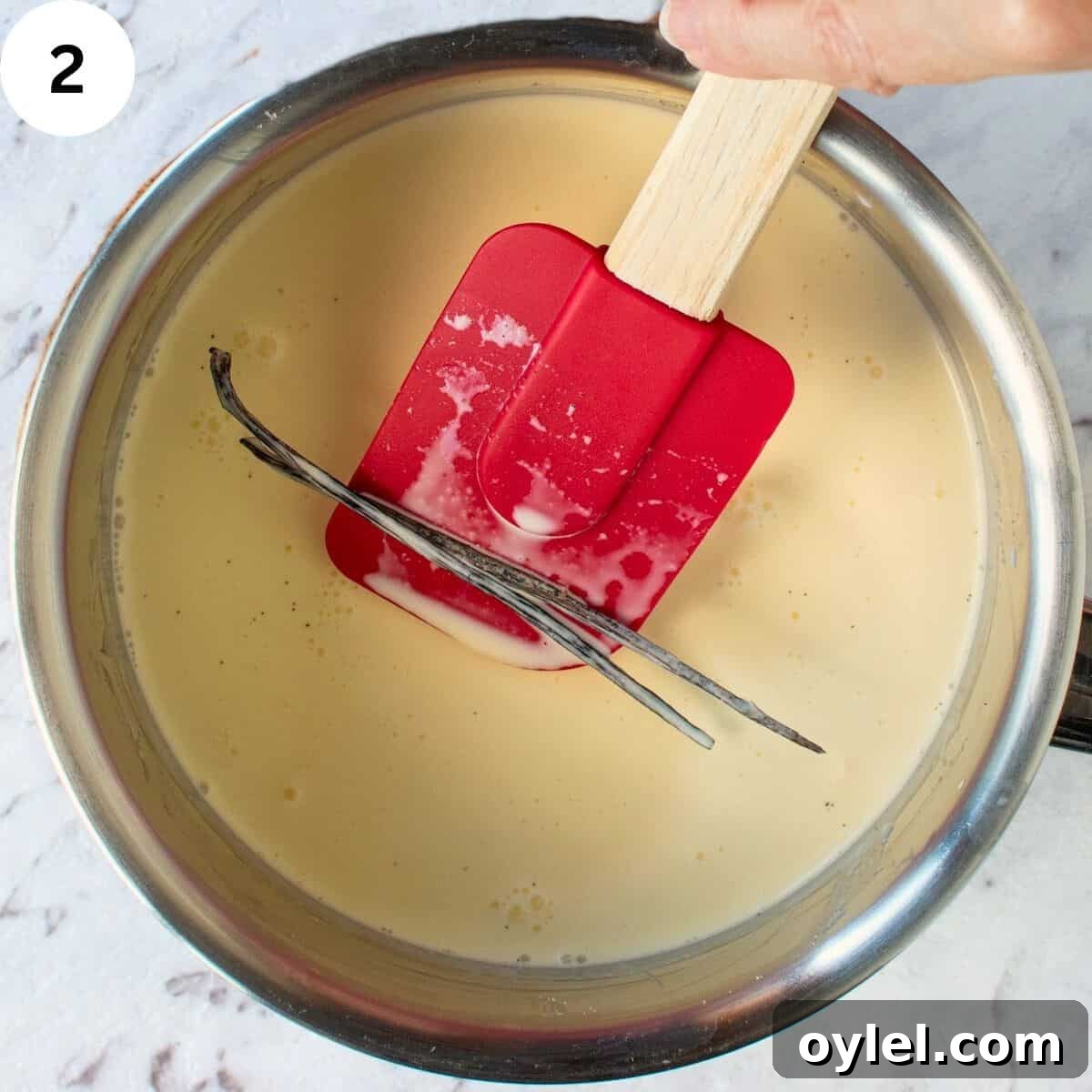 A split vanilla bean being lifted with a silicone spatula in a saucepan of cream.