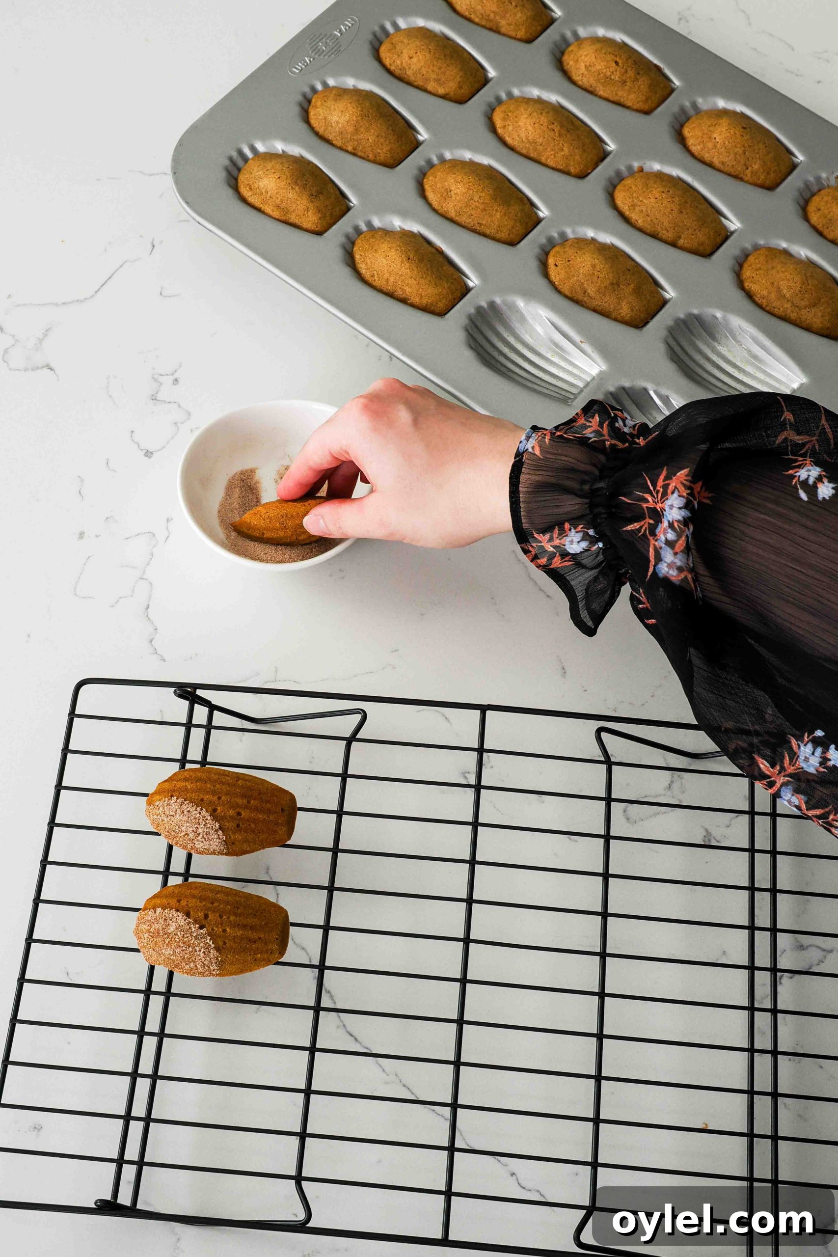 A hand dips a warm pumpkin madeleine into a small bowl of cinnamon sugar.