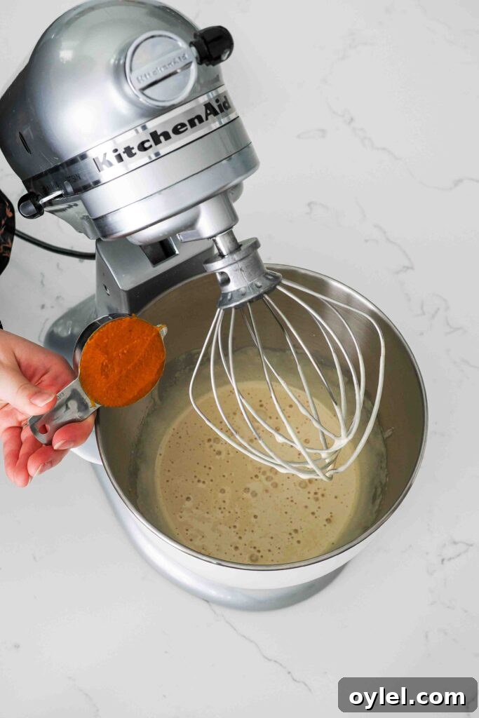 A hand holds a measuring cup with pumpkin over the bowl of a stand mixer.