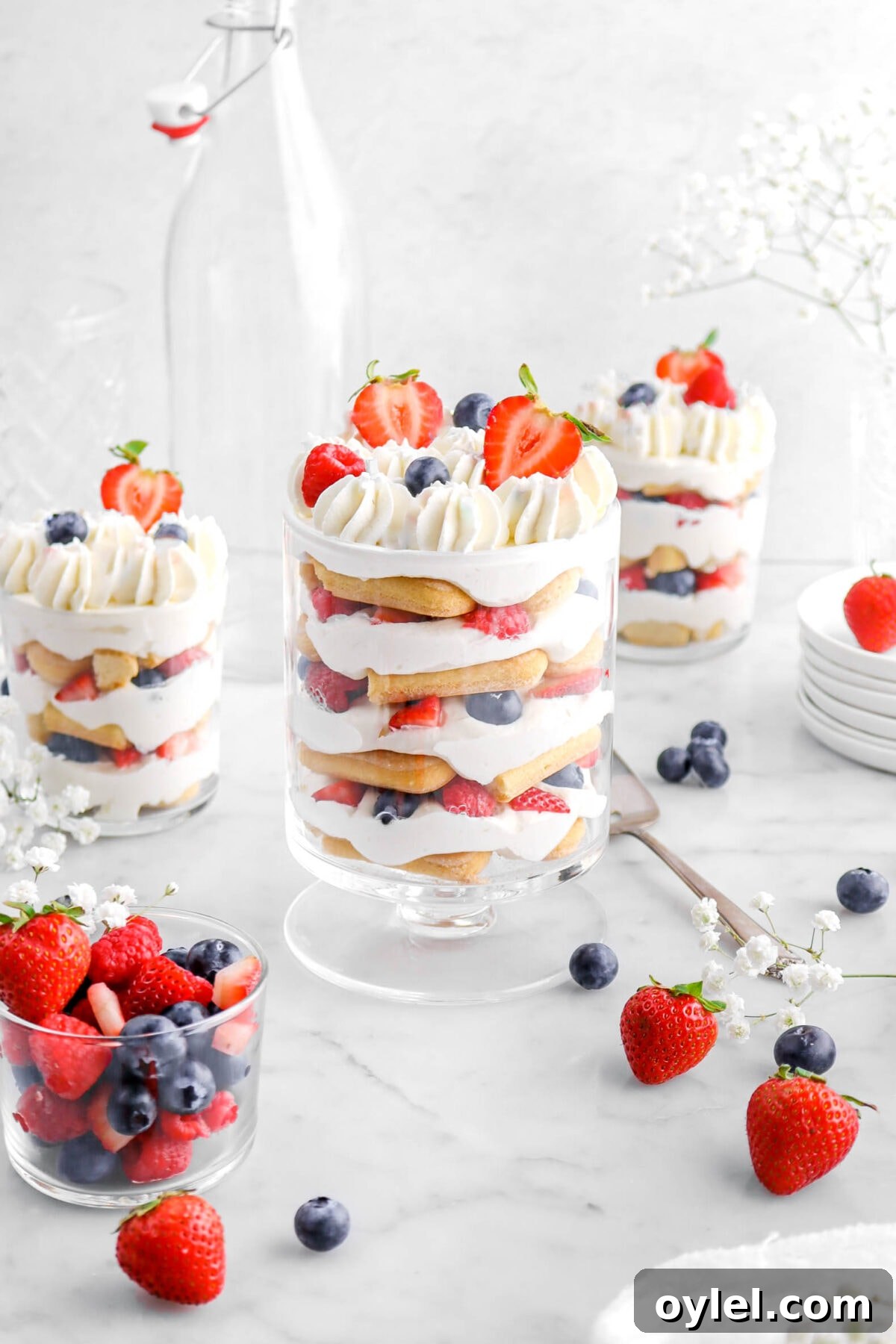 No-Bake Summer Berry and Ladyfinger Trifle Recipe 13 mixed berry trifle in tall trifle glass with two smaller glasses of berry trifle behind, with fresh berries and white flowers around, two empty glasses behind, and a stack of plates beside.