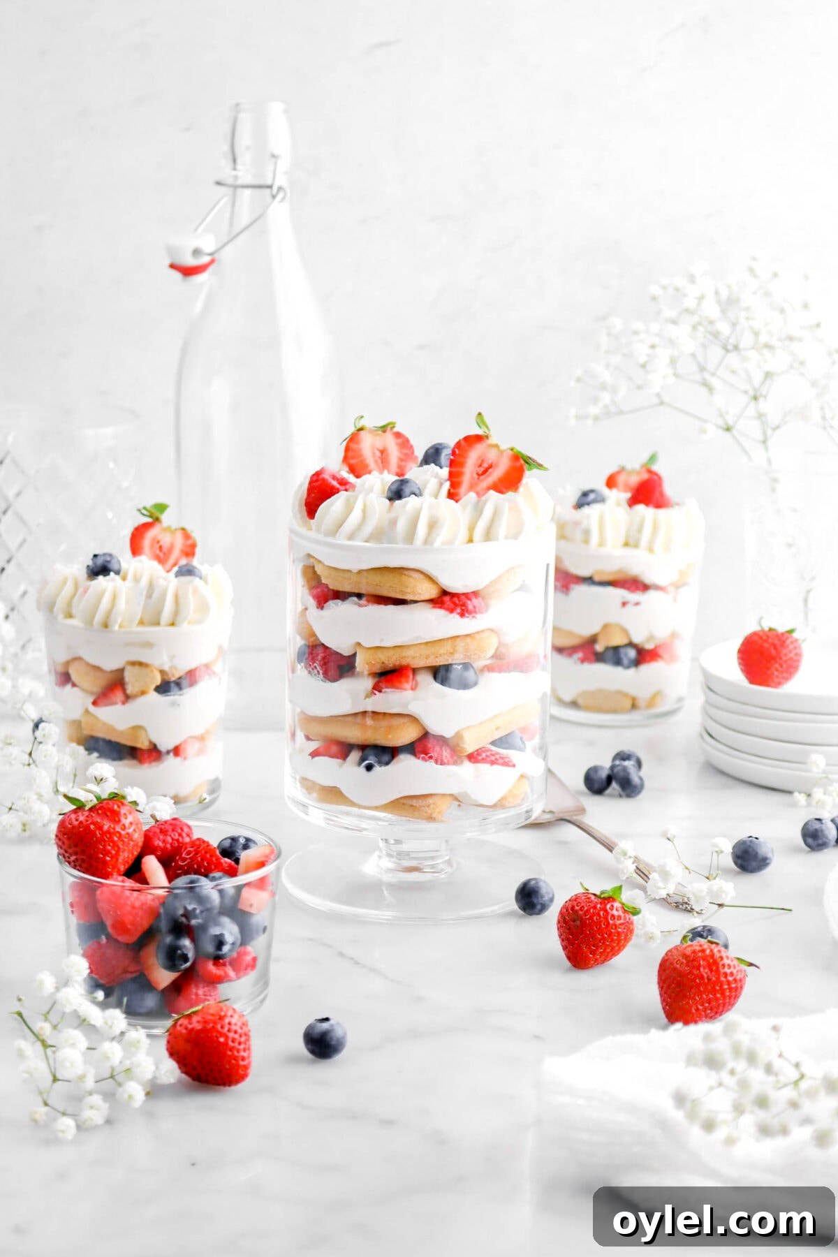 No-Bake Summer Berry and Ladyfinger Trifle Recipe 3 front shot of three berry trifles on marble surface with a glass of berries beside, fresh berries scattered around marble surface with white flowers around, two empty glasses behind trifle, and stack of plates beside.
