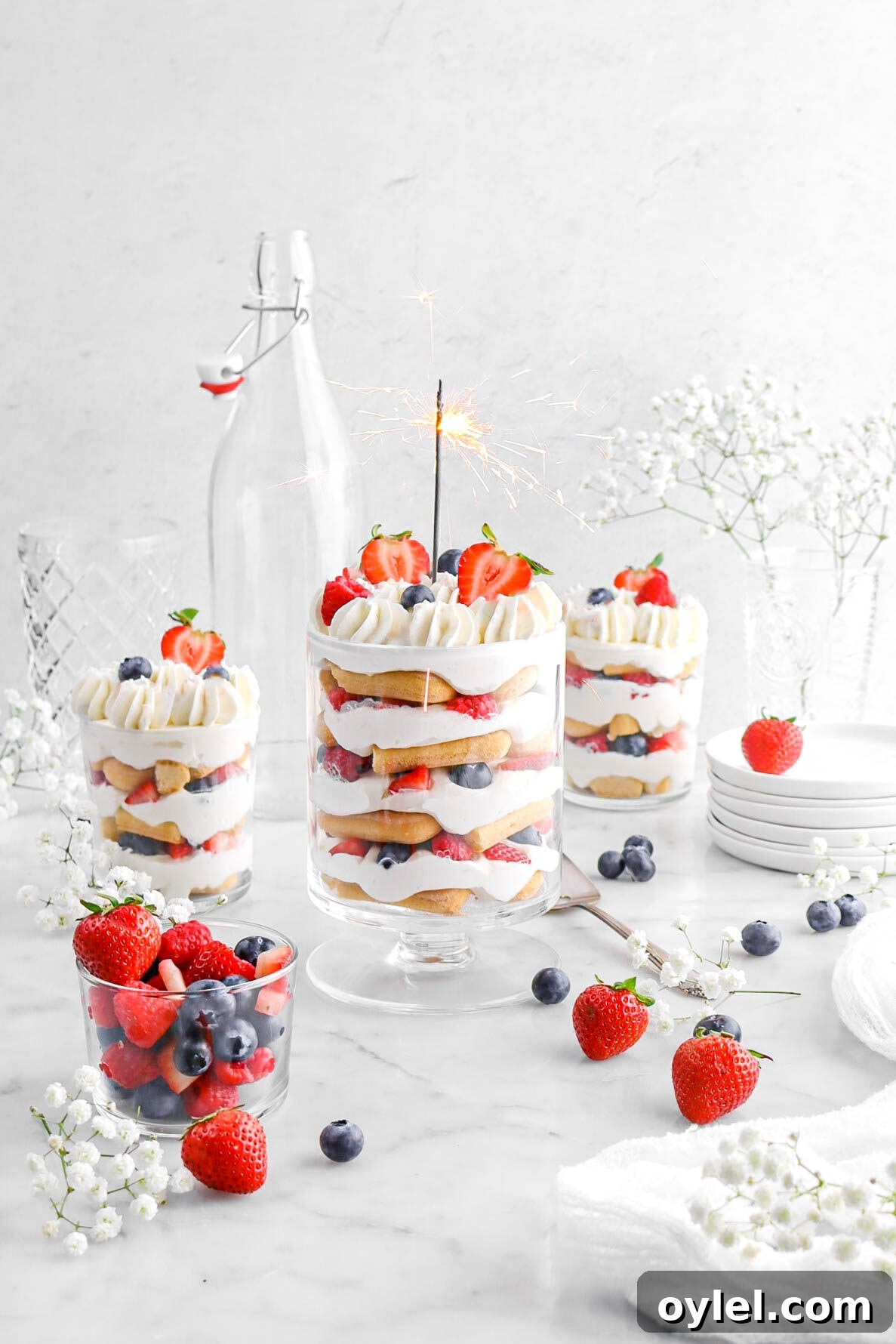 No-Bake Summer Berry and Ladyfinger Trifle Recipe 4 berry trifle with lit sparkler in main trifle, with two more smaller trifles behind, with white flowers and berries around.