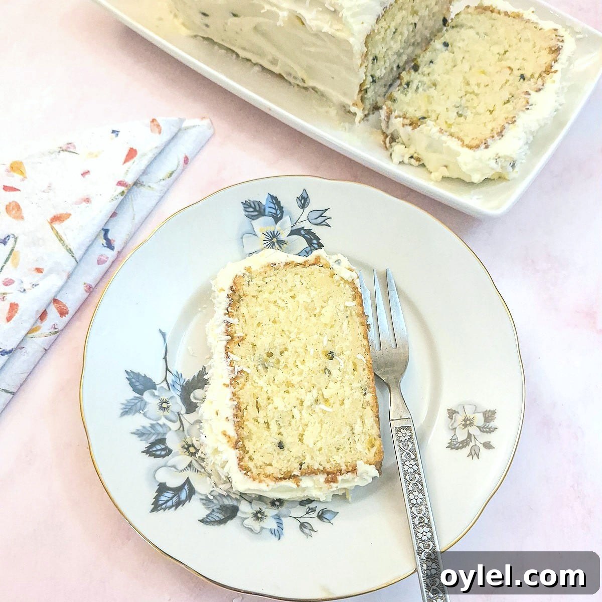 A slice of granadilla coconut loaf cake with a coating of cream cheese frosting on a small blue-flowered patterned plate.
